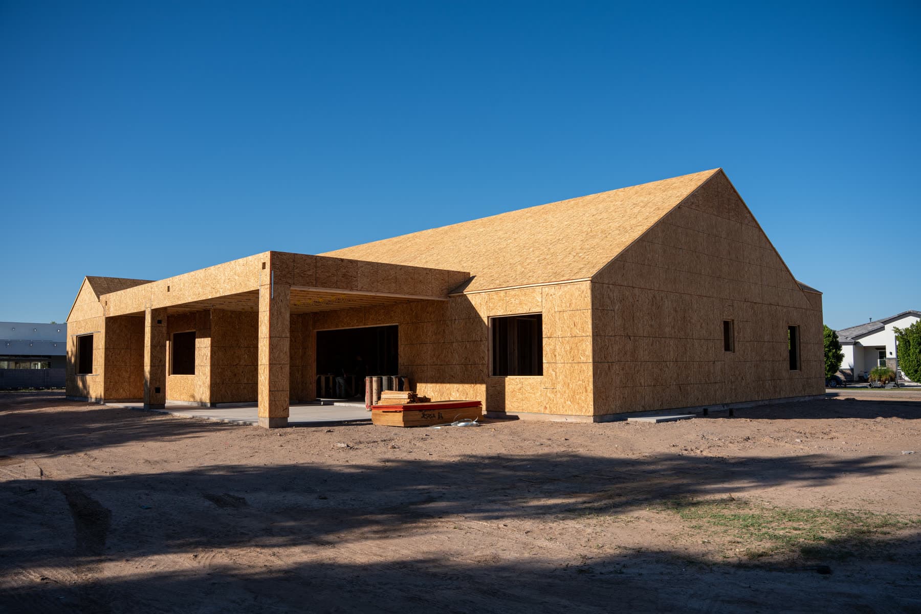 A framed and sheathed custom home on a dirt lot under a clear sky