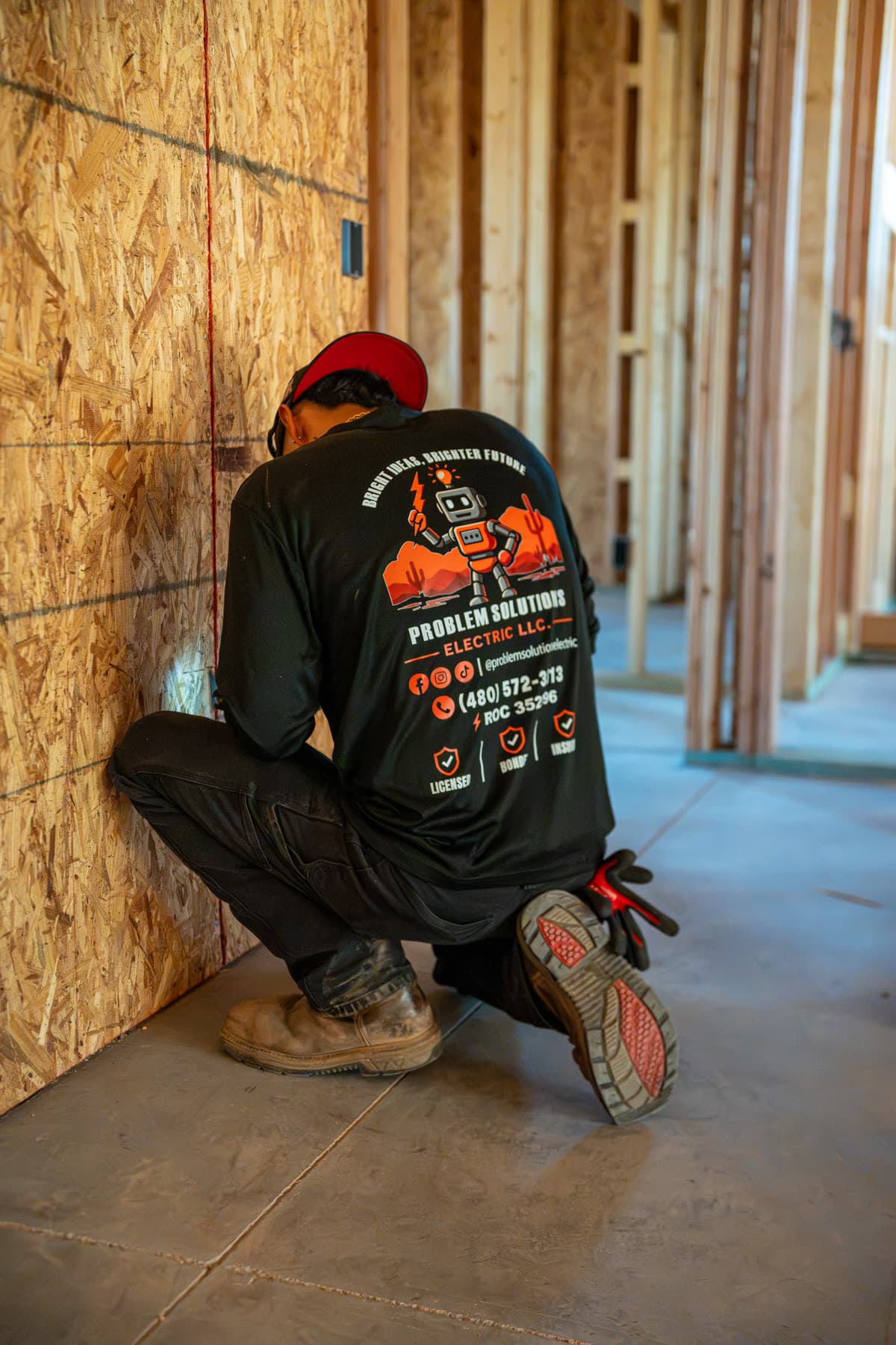 A technician in a company shirt kneeling at a framed wall during rough-in