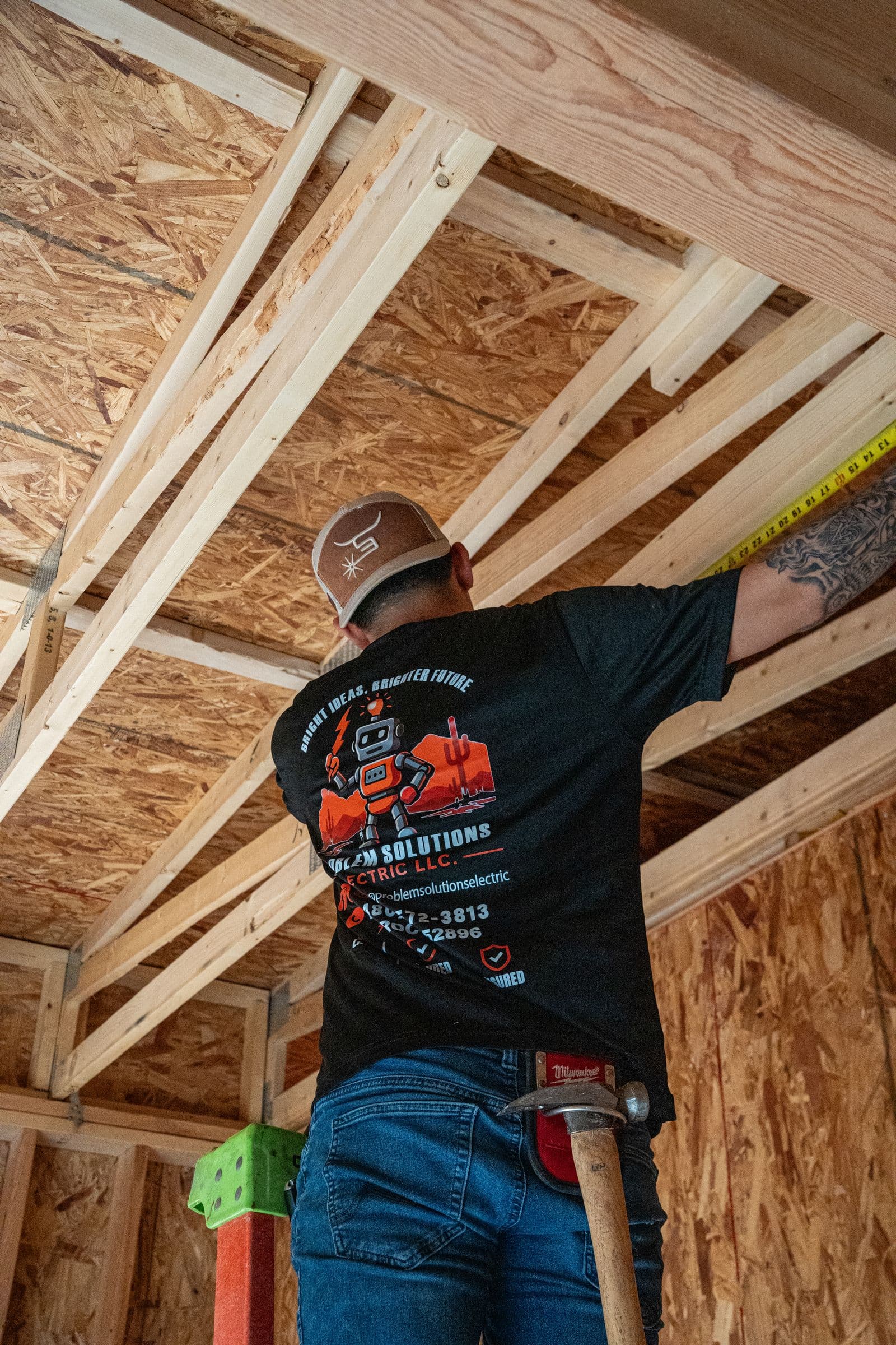A technician in a company robot shirt measuring between roof trusses with a tape