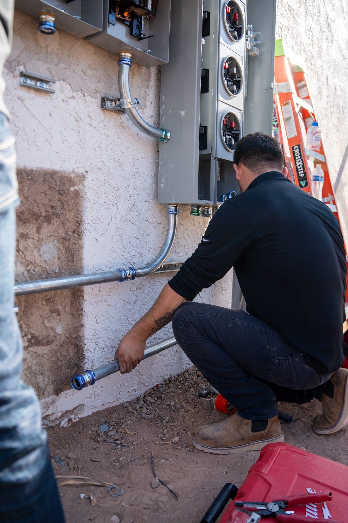A technician kneeling to fit conduit along a stucco wall below an electrical panel