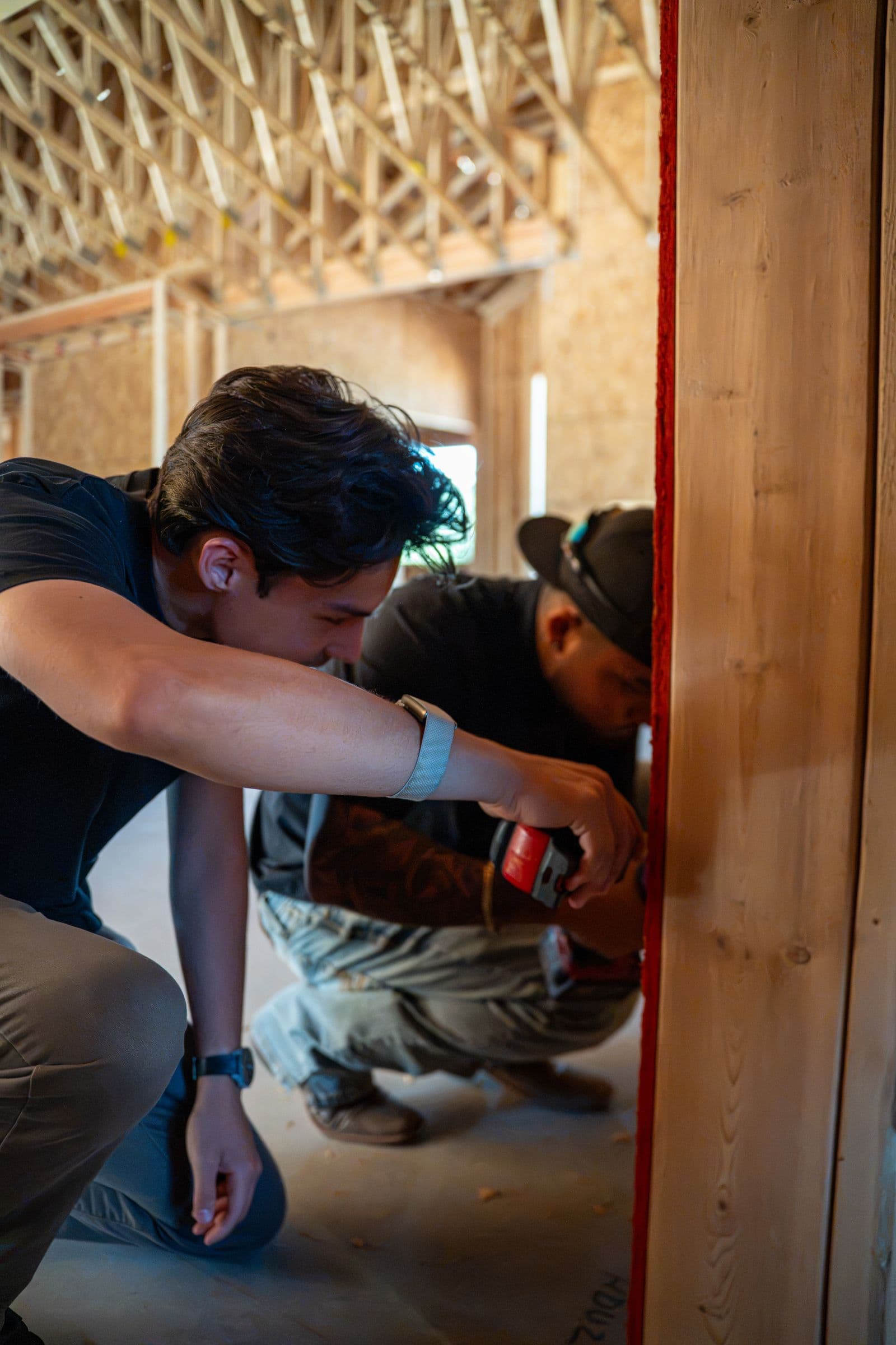 Two technicians crouched at a stud, one drilling a hole to run wire